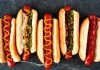 Dünya mutfağında geniş bir yer edinen hot dog, özellikle Amerika Birleşik Devletleri’nde adeta bir sembol haline gelmiştir. Kökeni 19. yüzyılın sonlarına kadar uzanan bu yiyecek, Almanya’dan Amerika’ya göç edenlerin beraberinde getirdiği sosis kültürü ile ortaya çıkmıştır. Uzun ekmek arasına yerleştirilen sosisin seneler içinde farklı soslarla buluşması, hot dog’u bugünkü popüler formuna kavuşturmuştur. Konulu bir haber görseli.