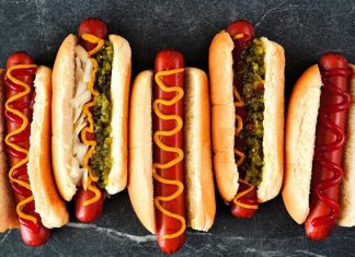 Dünya mutfağında geniş bir yer edinen hot dog, özellikle Amerika Birleşik Devletleri’nde adeta bir sembol haline gelmiştir. Kökeni 19. yüzyılın sonlarına kadar uzanan bu yiyecek, Almanya’dan Amerika’ya göç edenlerin beraberinde getirdiği sosis kültürü ile ortaya çıkmıştır. Uzun ekmek arasına yerleştirilen sosisin seneler içinde farklı soslarla buluşması, hot dog’u bugünkü popüler formuna kavuşturmuştur. Konulu bir haber görseli.