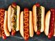 Hot Dog: Sokak Lezzetlerinin Vazgeçilmezi Dünya mutfağında geniş bir yer edinen hot dog, özellikle Amerika Birleşik Devletleri’nde adeta bir sembol haline gelmiştir. Kökeni 19. yüzyılın sonlarına kadar uzanan bu yiyecek, Almanya’dan Amerika’ya göç edenlerin beraberinde getirdiği sosis kültürü ile ortaya çıkmıştır. Uzun ekmek arasına yerleştirilen sosisin seneler içinde farklı soslarla buluşması, hot dog’u bugünkü popüler formuna kavuşturmuştur. Konulu bir haber görseli.